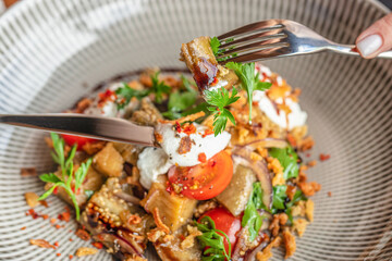 Close-up of a fork picking up roasted eggplant salad with cherry tomatoes, cream cheese, herbs, and...