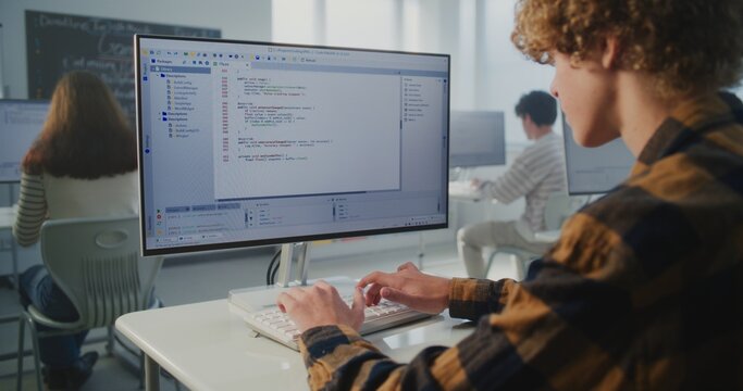 Young Student Sits at Desk in Modern Bright Classroom, Focused on Computer Screen While Typing Programming Code. Concept of Modern Education, Computer Science Learning, and Future Technology Skills. - Powered by Adobe