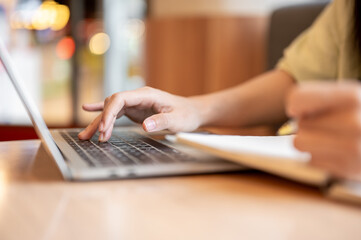 Close up of a woman student's hand typing on laptop aside notebook on wooden table in the cafe.