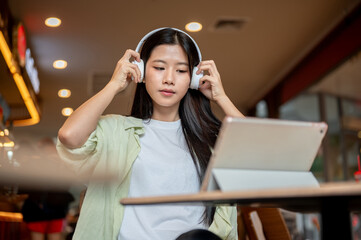 Pretty asian woman student wearing headphones while looking at tablet on wooden table in the cafe.