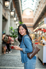 Smiling asian woman in denim holding phone while standing sideway in shopping mall or indoor market.