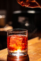 Close-up of a Negroni cocktail with ice cube on a wooden bar counter, illuminated by warm light