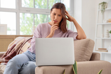 Naklejka premium Beautiful young thoughtful woman with laptop sitting on sofa in living room at home