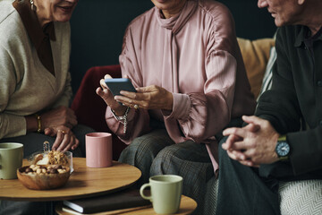 Three middle aged Caucasian women and man sitting together sharing smartphone screen, engaging in conversation, hands visible holding device, coffee mugs and snacks on table