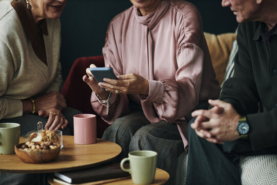 Three middle aged Caucasian women and man sitting together sharing smartphone screen, engaging in conversation, hands visible holding device, coffee mugs and snacks on table
