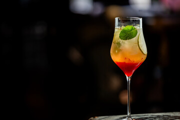 Layered cocktail in a stemmed glass with red, orange, and yellow hues, garnished with mint and lime slice. Dark background highlights vibrant drink colors. Vertical, close-up, studio shot.