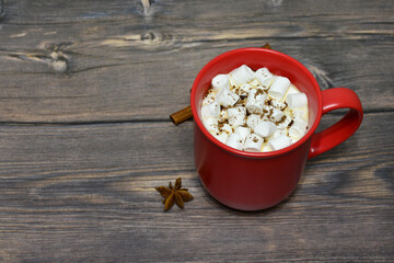 Cozy red mug of hot chocolate with marshmallows, cinnamon and star anise on a wooden surface