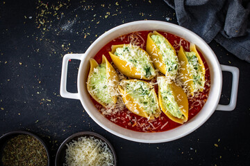 Pasta shells stuffed with cheese and spinach in tomato sauce in baking dish prepared for baking on black table. Top view