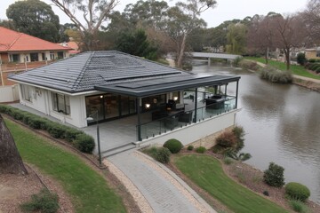 Modern riverside house with glass-railed terrace and outdoor seating overlooking a calm river, manicured lawns, trees, and a small pedestrian bridge.