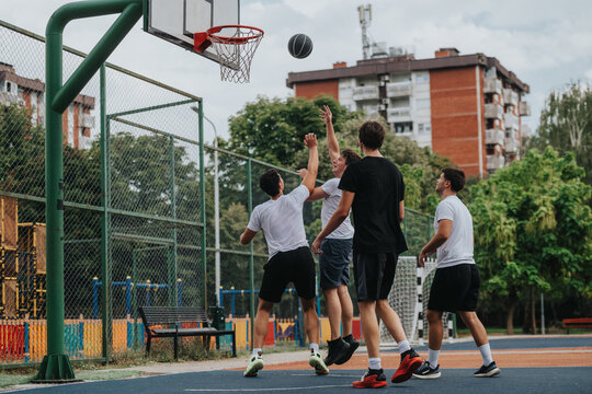A group of young men compete in a fast-paced pickup basketball game on an open fenced court in a city park, wearing white and black athletic outfits as they reach for the ball. - Powered by Adobe