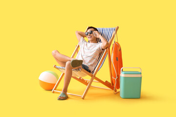 Young Asian man talking by mobile phone on deckchair against yellow background