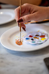 Hand mixing paint with brush on white plate and colorful palette