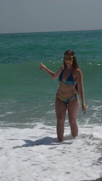 Woman, beach, ocean, young woman in bikini with golden mask stands in turquoise waves. Vertical video.
