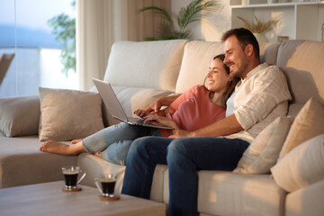 Happy couple at home using laptop on a couch at sunset