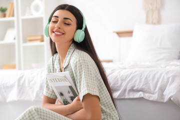 Beautiful young woman with modern headphones and magazine listening to music on floor in bedroom