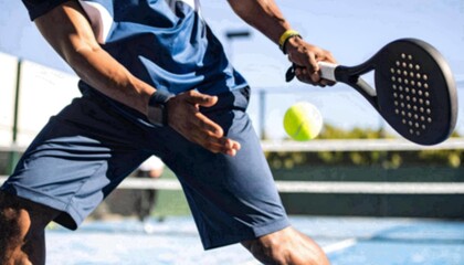 African American male padel player in dark sports attire making a forehand or low volley shot. He is shown mid-swing, hitting the yellow ball with a black padel racket on an outdoor court.