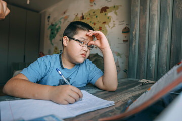 Asian boy with glasses holding head in frustration, school task at home desk near world map wallpaper, concept of homeschooling, education service, tutoring support