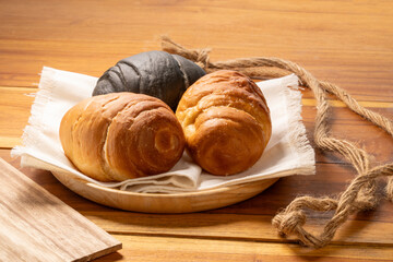 Three round croissants, one dark charcoal and two classic golden in wood plate on wooden table background, Round croissants or Croissant Buns in wood plate.