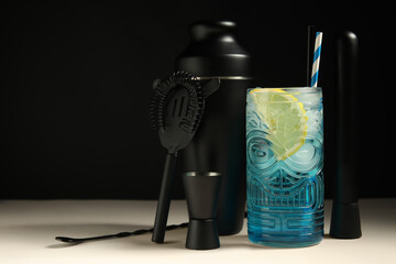 Glass of fresh cocktail and bartender tools on white table near black wall, closeup