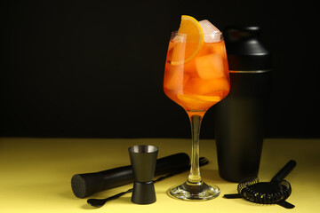 Glass of Aperol spritz cocktail and bartender tools on yellow table near black wall, closeup