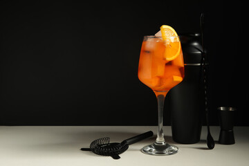 Glass of Aperol spritz cocktail and bartender tools on white table near black wall, closeup