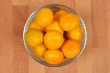 An overhead shot shows a clear glass bowl filled with small, round, bright yellow and orange mandarins resting directly on a light wooden surface