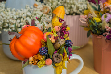  close up of an autumn themed floral arrangement placed in a white mug, featuring a small pumpkin, ceramic mushroom, dark berries, dried flowers, and nuts, concept of fall decor and thanksgiving
