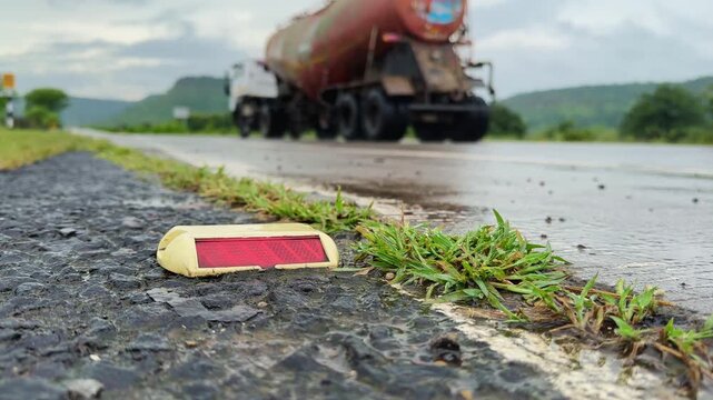 Static close-up of a wet road reflector marker on asphalt after rain, with a blurred truck driving away in the background on a countryside highway.