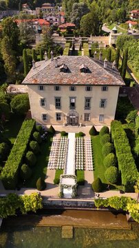 Aerial drone view of Villa Balbiano on Lake Como during a luxury outdoor wedding with floral arch and rows of white chairs on green lawn, overlooking panoramic lake and mountain views