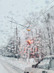 The view from inside a car driving through a city street during snowfall, passing a park with snow-covered trees.
