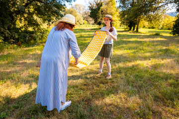 Mother and daughter preparing picnic area by laying blanket on lawn in natural park. Concept of family summer rest and active vacation outdoors.