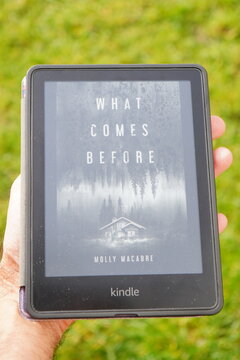 A person holds an Amazon Kindle e-reader displaying the cover of the book 'What Comes Before' by Holly Macabre outdoors