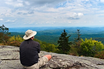 A solo hiker, in the Blue Ridge Mountains, sits on a rocky overlook and enjoys looking out over the valley after hiking to the top of a trail.