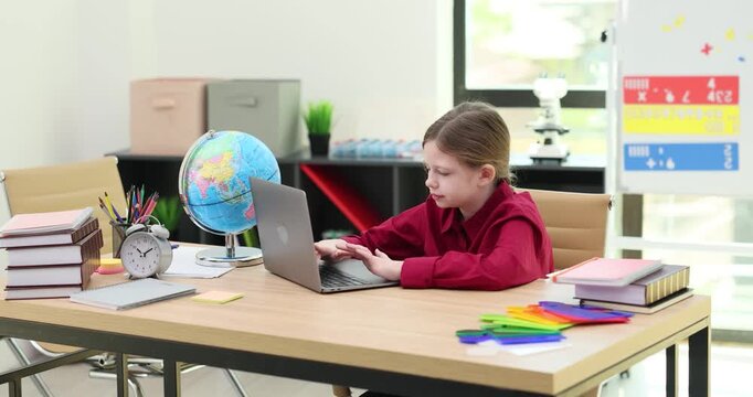 Focused little schoolgirl types on laptop keyboard while sits at desk. Girl student does online tests on notebook computer learning online at home