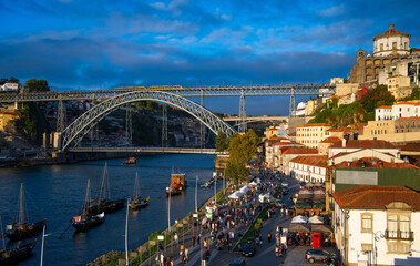 Fototapeta premium Panoramic evening view of the old town of Porto and Gaia in the Douro Valley (Portugal). Waterfront promenade with boats and numerous tourists and the iconic double-decker bridge in the background.