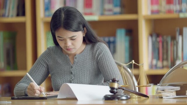 Asian female law student focused on studying with tablet, legal scale, and gavel on desk in university library.
