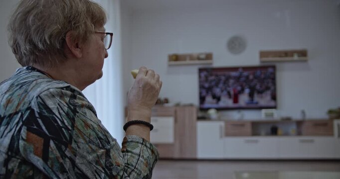Elderly woman sitting on a sofa in her living room, eating a snack and watching TV. She's wearing glasses and a patterned blouse.