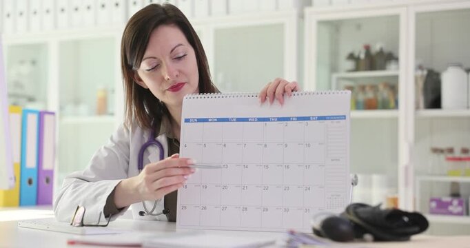 Woman doctor in white lab coat holds up calendar and points at free date. Physician shows available slot for appointment in office. Slow motion