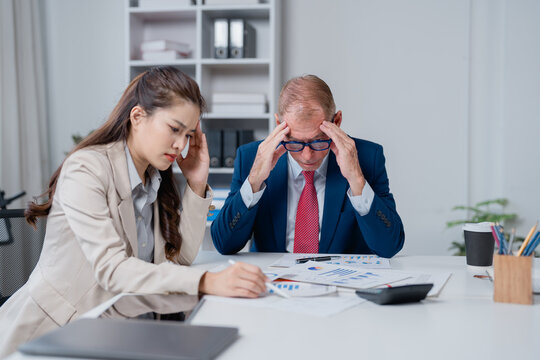 Asian businesswoman and senior male executive feeling overwhelmed and frustrated, suffering from headaches while reviewing financial documents and discussing business problems in a modern office