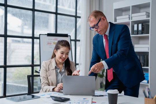 Experienced senior executive mentoring young female colleague. Pointing at laptop screen. Discussing business strategy. And analyzing financial data on documents in a collaborating office environment - Powered by Adobe