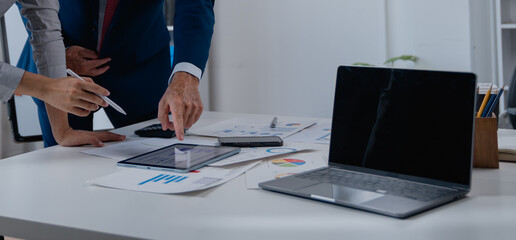 Business professionals collaborating at a desk, reviewing various charts, graphs, and reports with a tablet and laptop, symbolizing teamwork, financial analysis, and strategic planning