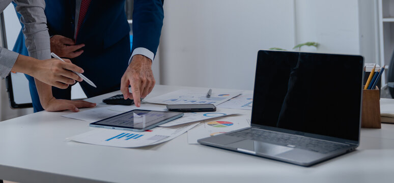 Business professionals collaborating at a desk, reviewing various charts, graphs, and reports with a tablet and laptop, symbolizing teamwork, financial analysis, and strategic planning