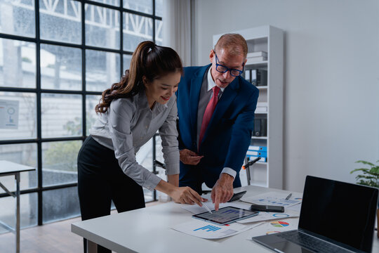 Business colleagues interacting in a modern office, a successful male executive mentoring a young female employee, analyzing financial data and charts on a digital tablet with documents