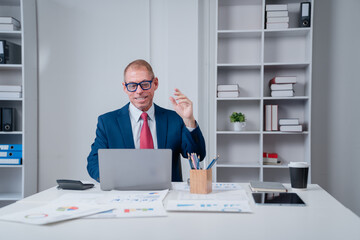 Businessman smiling and waving during a virtual meeting or online conference, sitting at his office desk with a laptop, documents, and coffee cup, focusing on communication and remote work