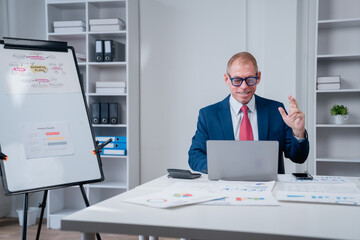 Senior businessman in a suit engaging in a video conference from his professional office, discussing business plans and reviewing charts and graphs on his desk