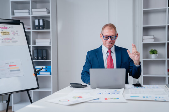 Smiling mature businessman in a professional suit and blue glasses communicating during an online video call or teleconference, sitting at an office desk with paperwork and a laptop