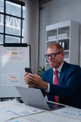 Mature businessman in a blue suit and glasses smiling while using a smartphone and laptop at his desk, surrounded by business documents and a whiteboard in a modern office