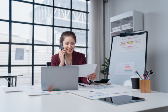 Asian businesswoman talking on a mobile phone while reviewing documents and using a laptop at a modern bright office desk, managing multiple tasks with a happy expression