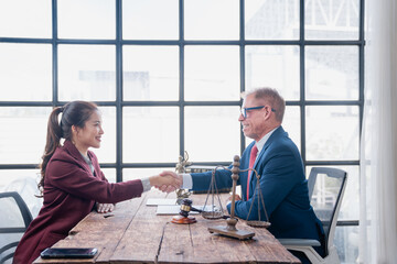 Experienced lawyer and young female client shaking hands across a rustic wooden desk in a modern office, symbolizing a successful legal consultation and agreement