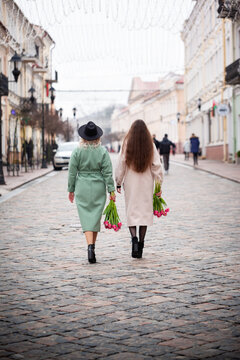 Two women walking hand in hand on a cobblestone street, dressed in stylish coats, one holding a bouquet of flowers, showcasing friendship and urban lifestyle. Backview